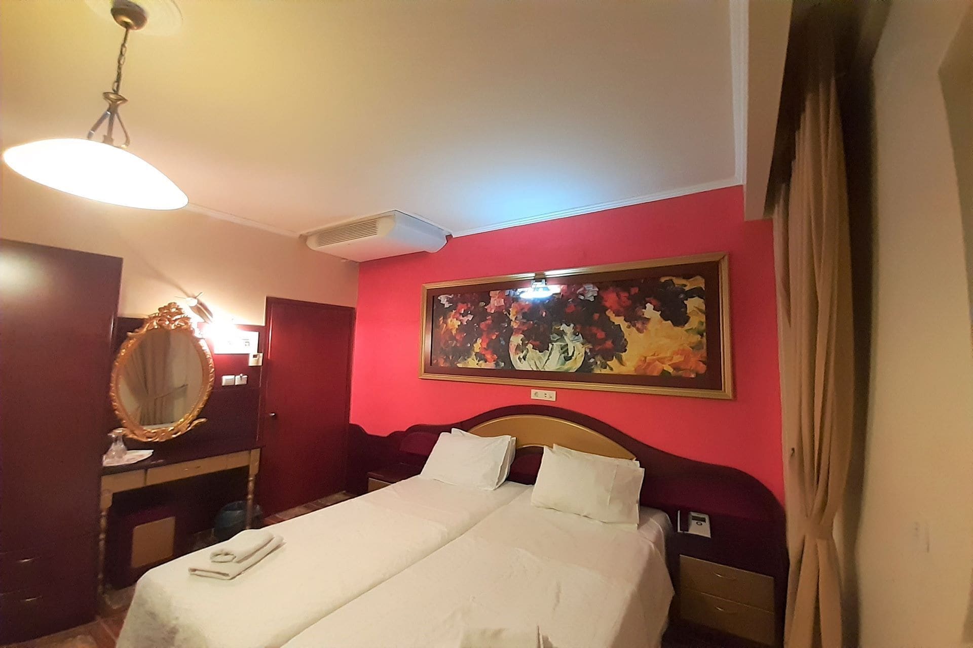 A hotel room with a bed, a mirror, a painting on the wall, and a ceiling light.