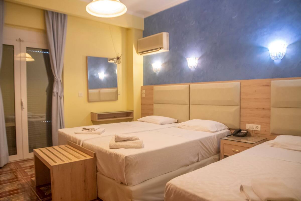 A hotel room with two beds, a wooden bench, a mirror, an air conditioning unit, and a telephone on a bedside table.
