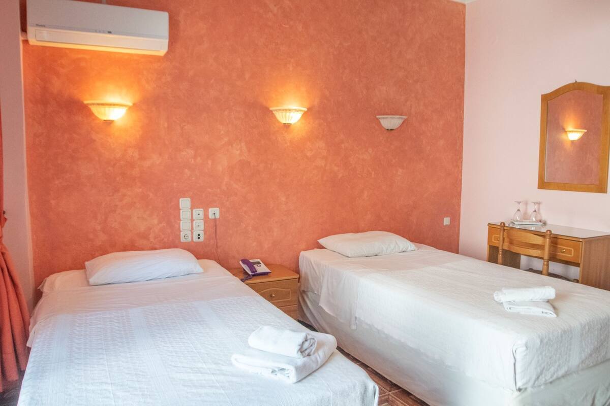 A hotel room with two single beds, a wooden nightstand, a desk with a mirror, and a wall-mounted air conditioner.