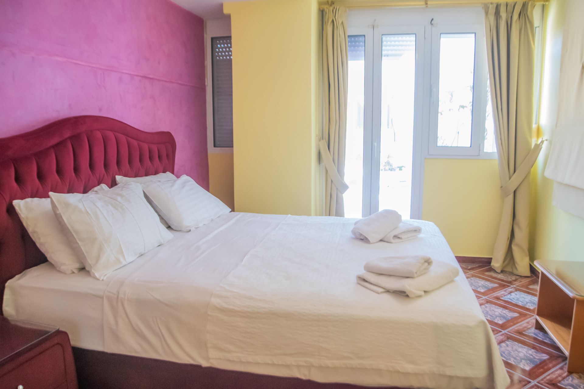 A hotel room with a bed, pillows, a window with curtains, and a tiled floor.