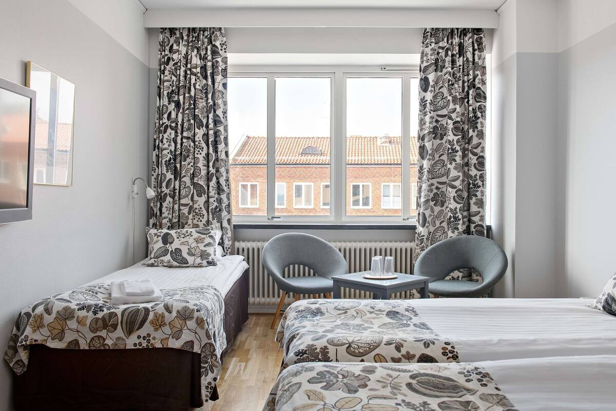 A hotel room with two beds, a small table, two chairs, and a window with curtains.