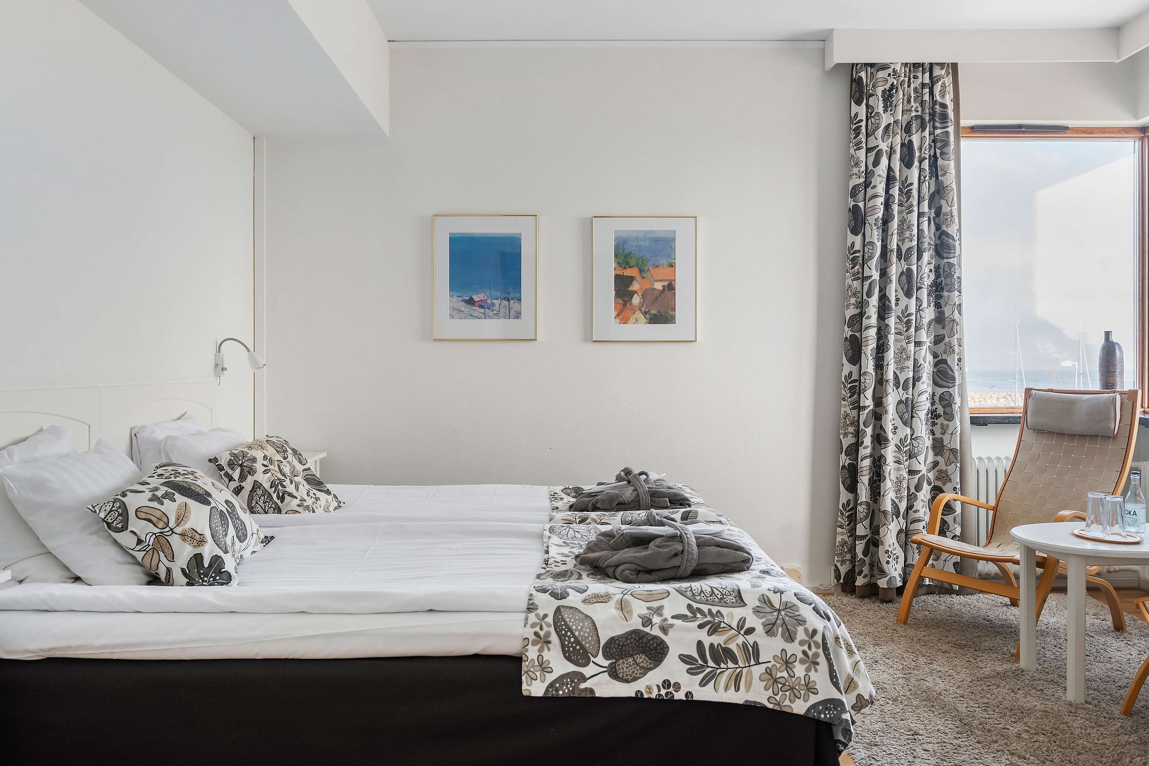 A hotel room with a bed, two framed pictures on the wall, a window with curtains, a chair, and a small table with a glass and a bottle.