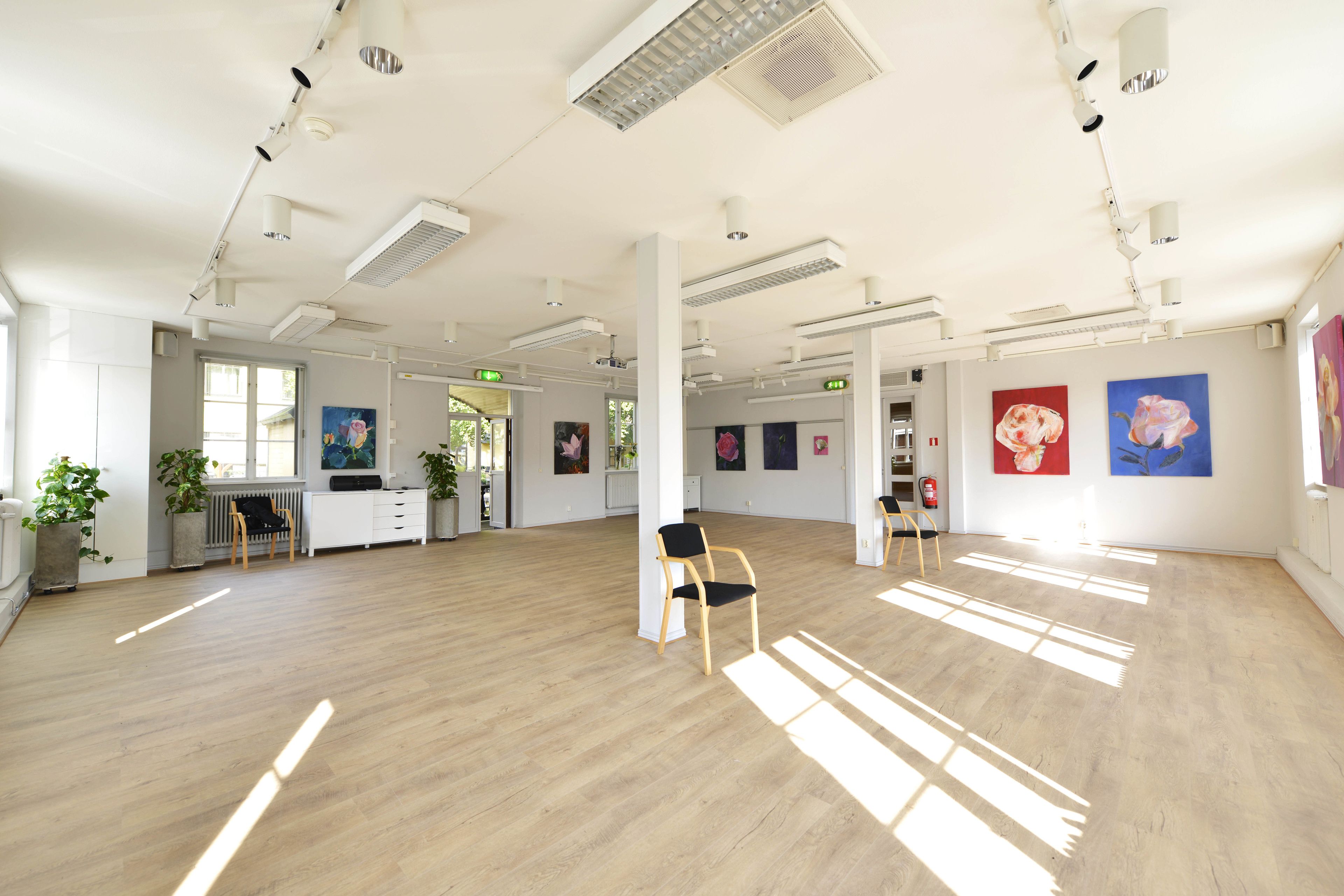 A spacious room with wooden flooring, white walls, and ceiling-mounted lighting. There are several chairs and a small table. Artwork is displayed on the walls.