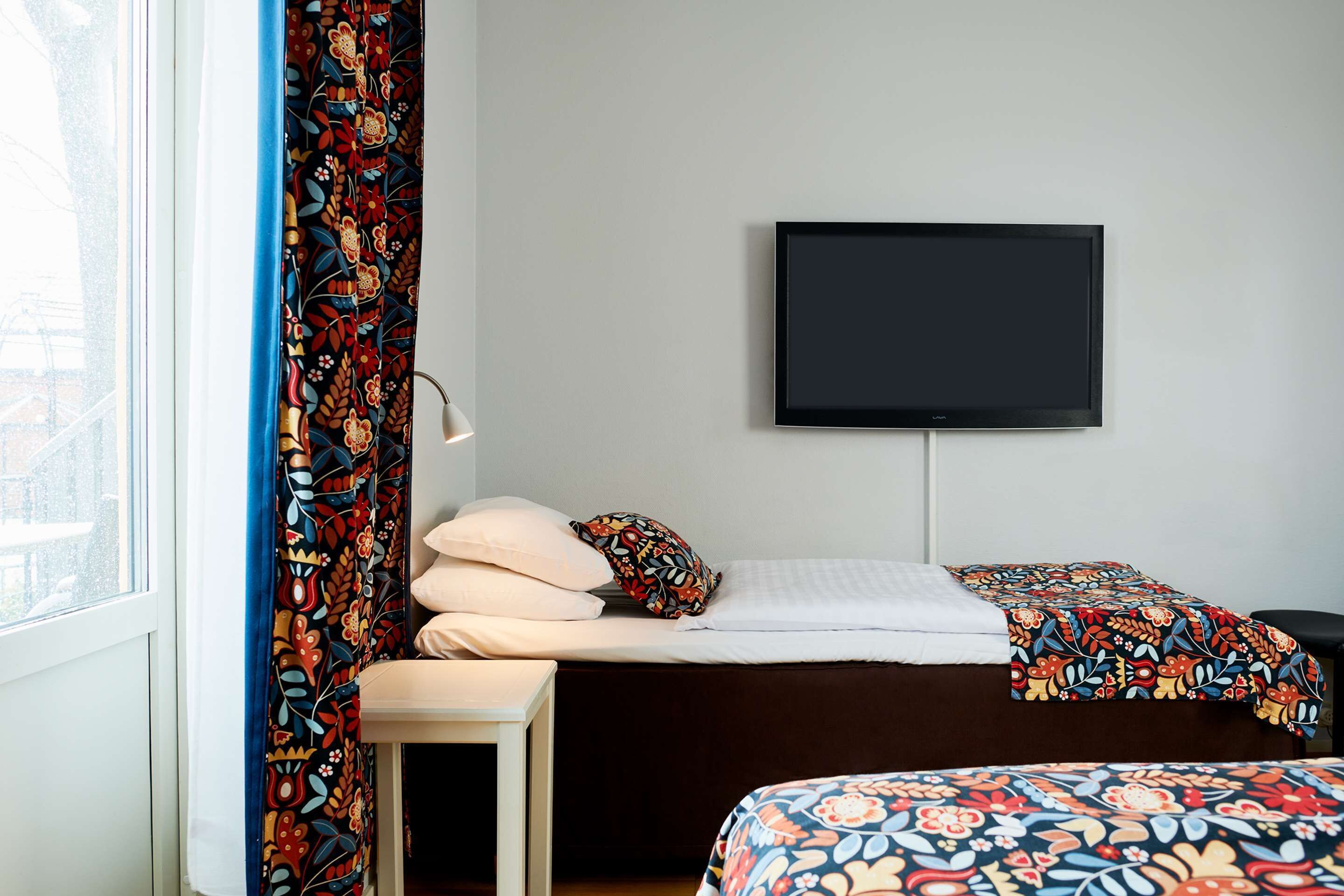 A hotel room with a bed, a TV mounted on the wall, a window with curtains, and a bedside table.