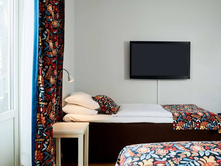 A hotel room with a bed, a TV mounted on the wall, a window with curtains, and a bedside table.