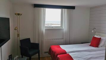 A hotel room with two beds, a desk with a chair, a floor lamp, and a view of the sea through a window.
