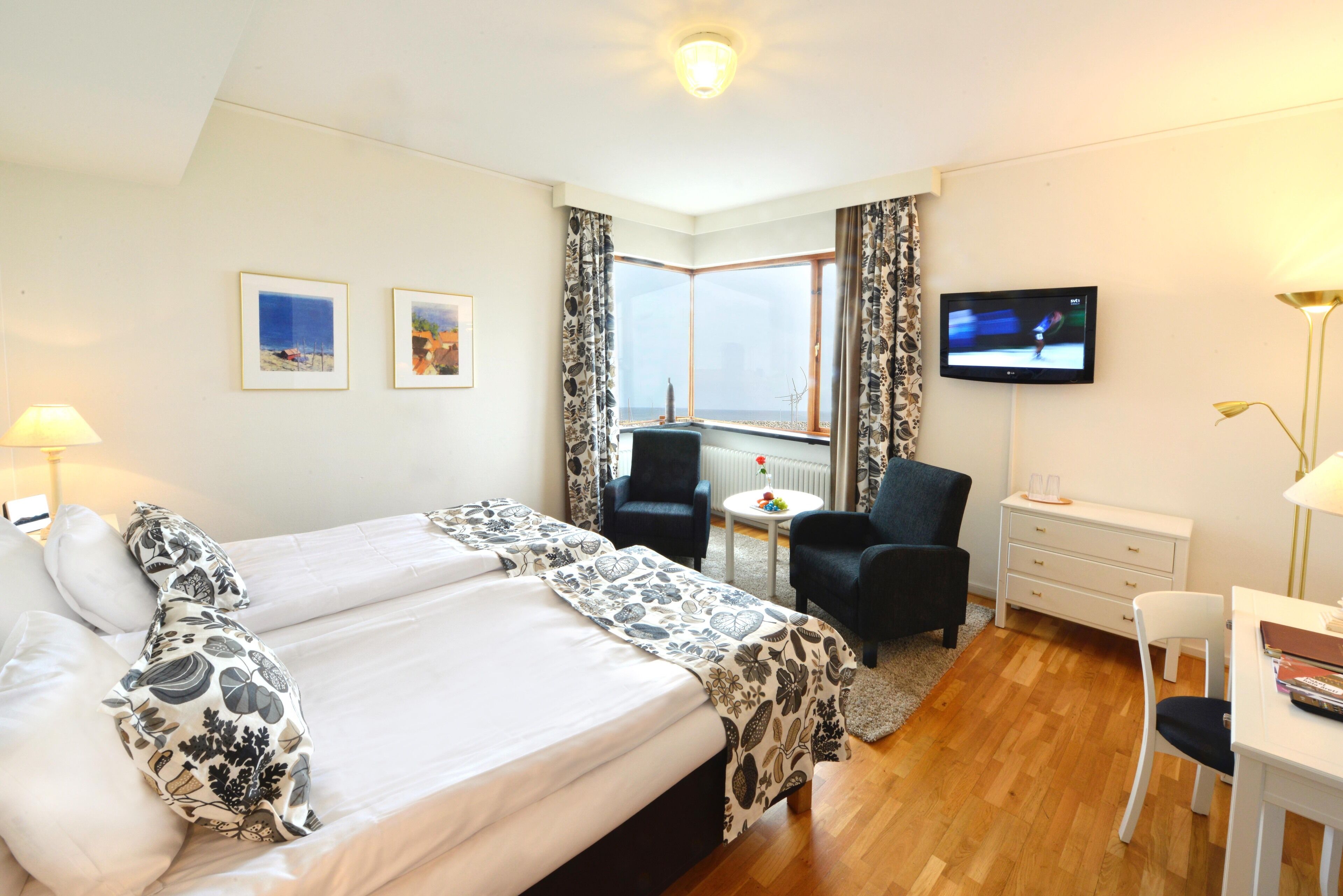 Ein Hotelzimmer mit einem großen Bett, zwei Stühlen, einem Schreibtisch, einem Fernseher und einem Fenster mit Vorhängen.
