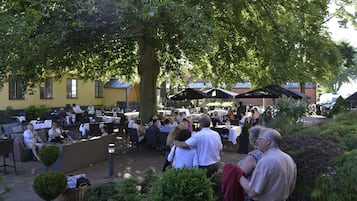 En udendørs café med borde og stole under et stort træ, omgivet af grønt og bygninger i baggrunden.