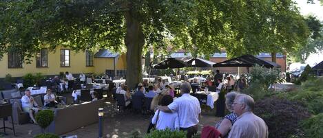 An outdoor cafe with tables and chairs under a large tree, surrounded by greenery and buildings in the background.