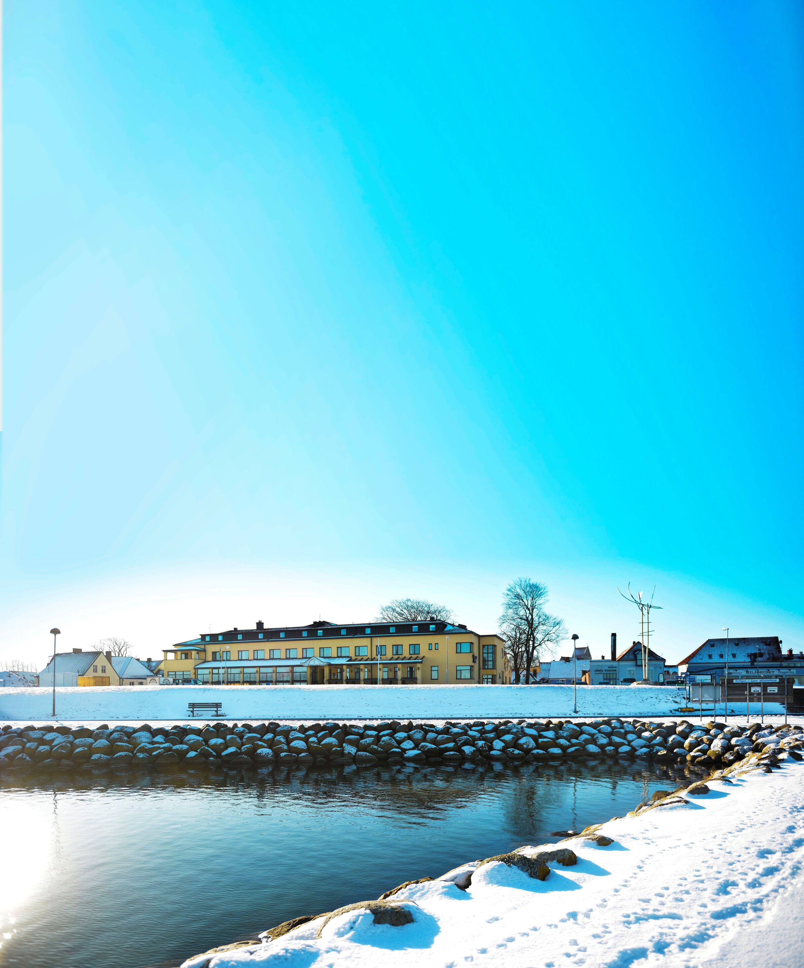 A snow-covered waterfront with a row of buildings, including a yellow building with multiple windows and a sign.