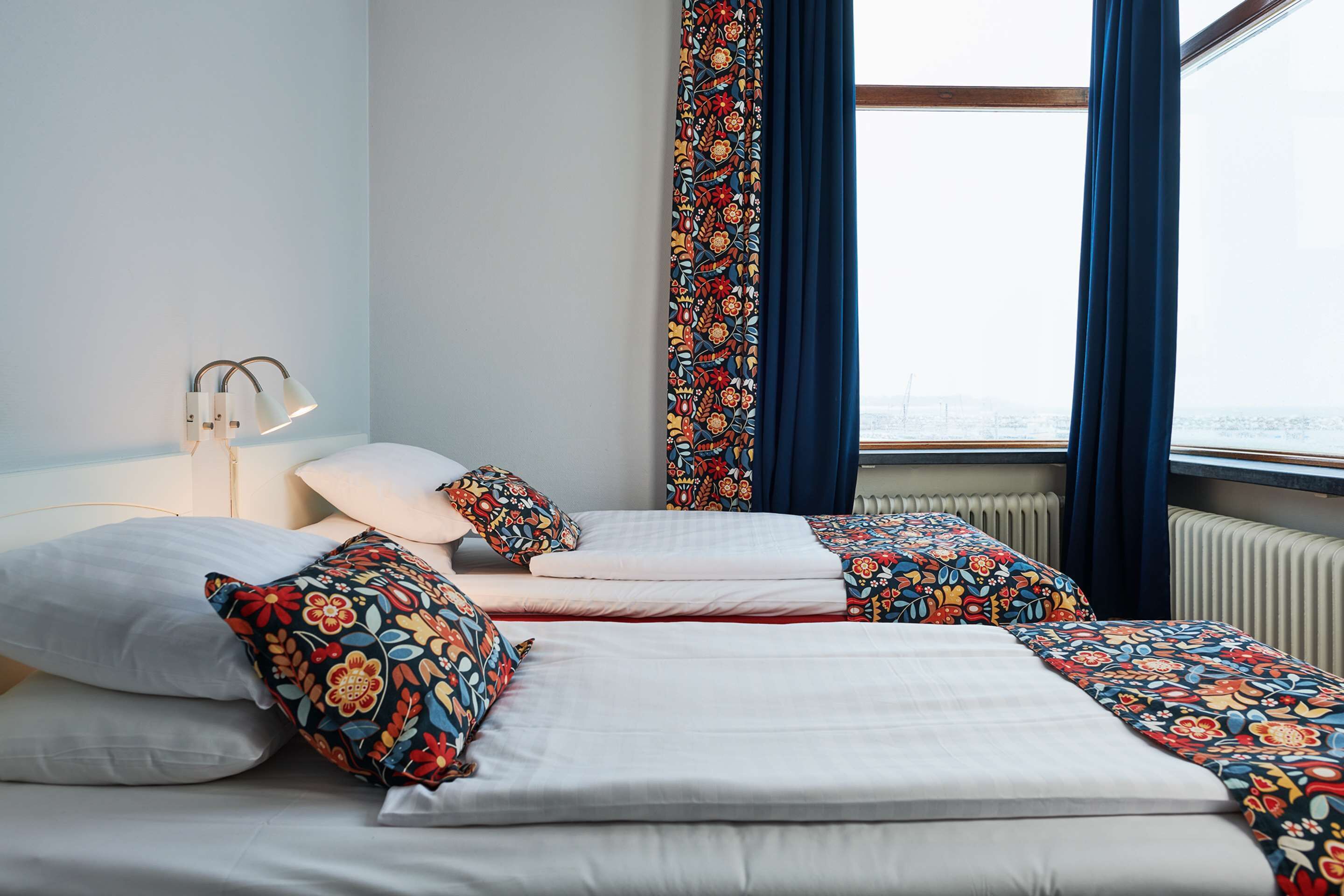 Une chambre d’hôtel avec deux lits, une table de chevet, une lampe et une vue sur la ville grâce à de grandes fenêtres.