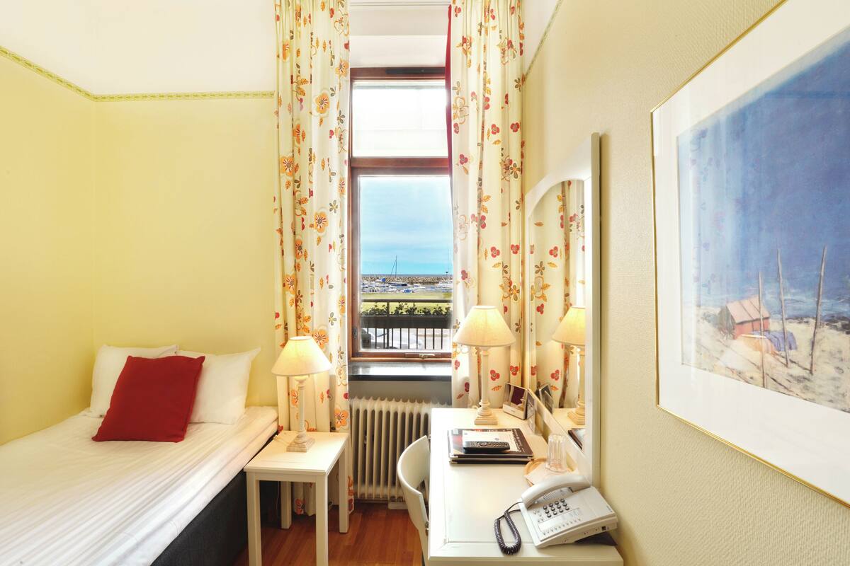 A hotel room with a bed, a desk with a lamp, a telephone, and a painting on the wall.