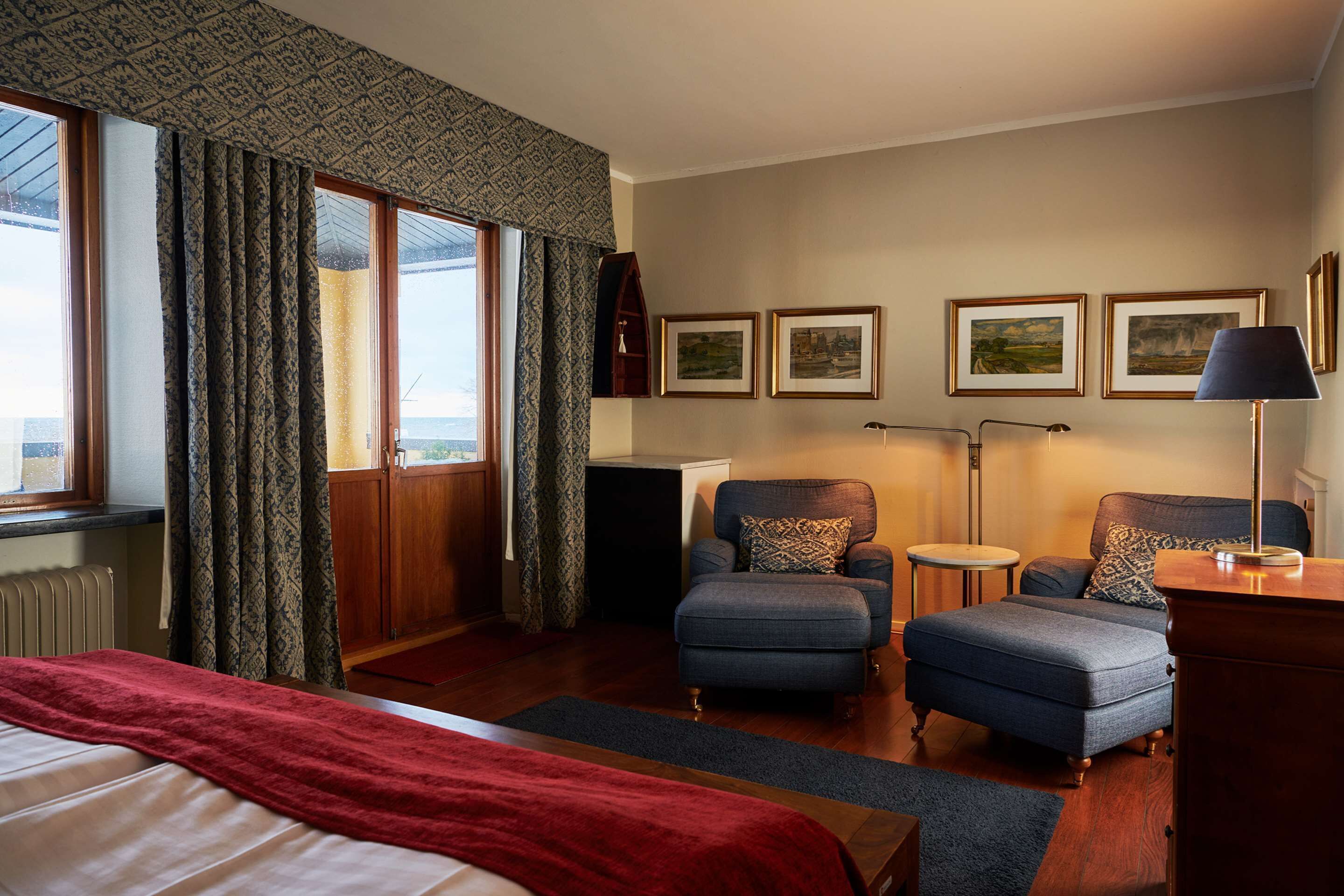 Ein Hotelzimmer mit einem Bett, zwei Sesseln, einem Nachttisch, einer Lampe und gerahmten Bildern an der Wand.