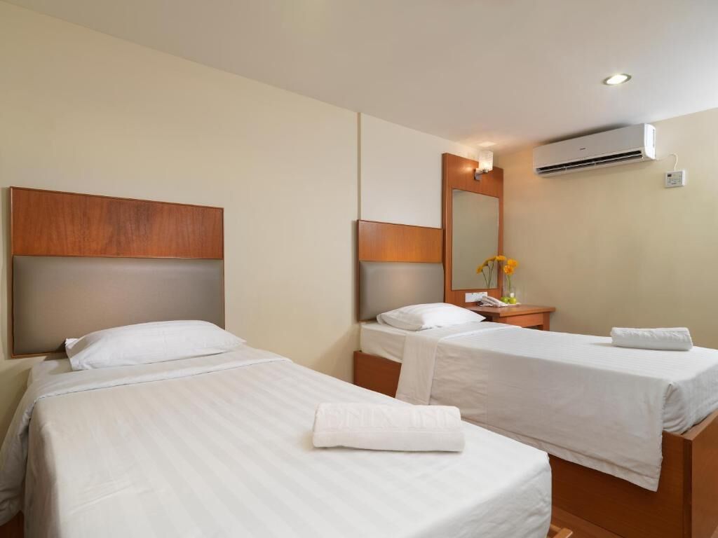 Habitación de hotel con dos camas individuales, cabecero de madera, mesita de noche con lámpara, espejo y aire acondicionado.