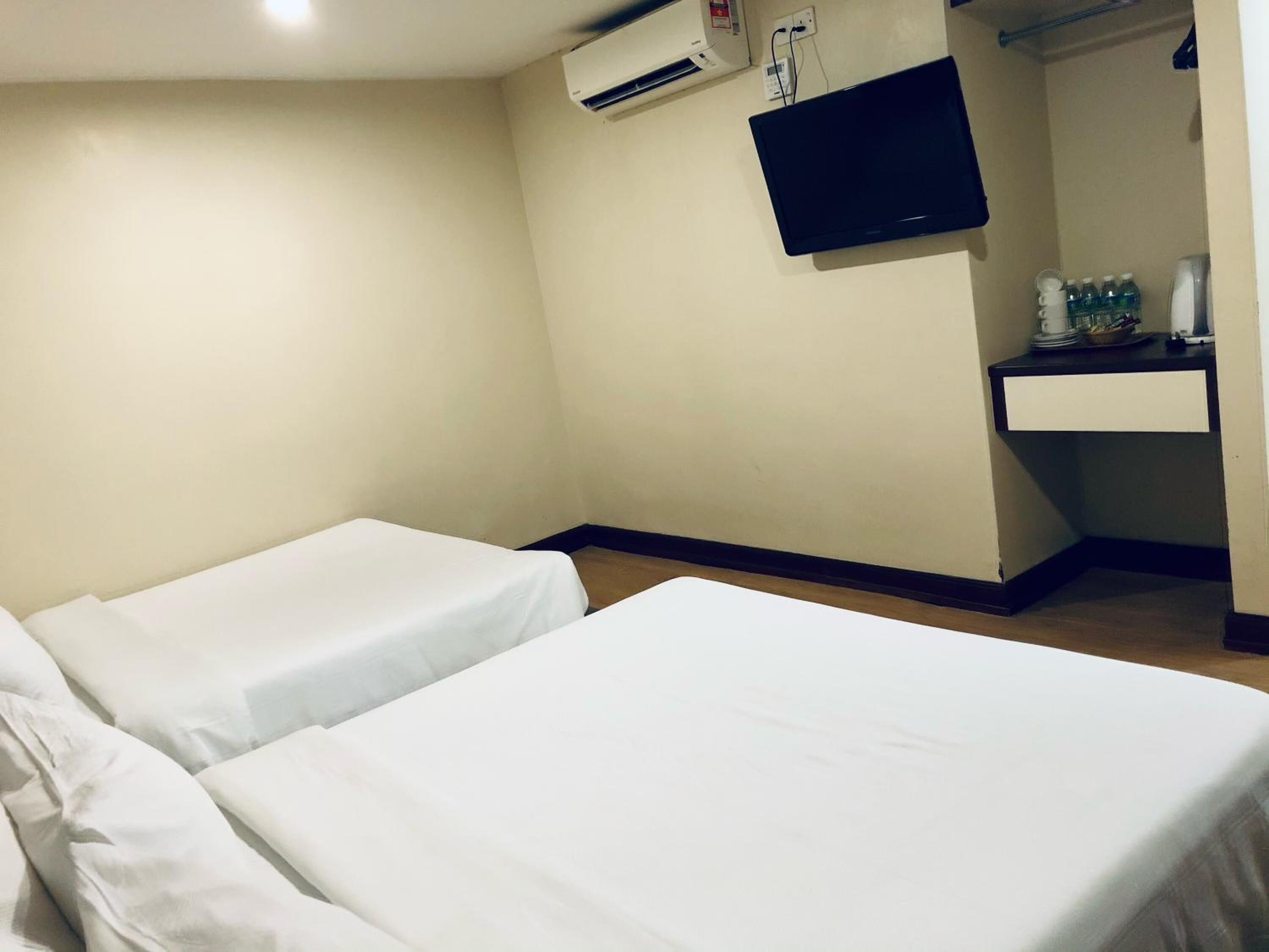 A hotel room with two single beds, a flat-screen TV mounted on the wall, a small desk with a chair, and a shelf with toiletries.