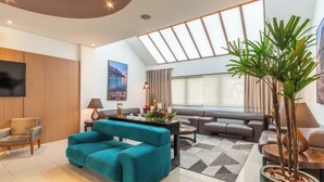A modern living room with a teal sofa, a grey sectional sofa, a coffee table, and a large window with a view of the outdoors. - Hotel Laghetto Viale (Gramado)