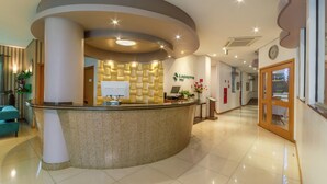 A reception area with a curved reception desk, a clock on the wall, and a staircase in the background. - Hotel Laghetto Viale (Gramado)