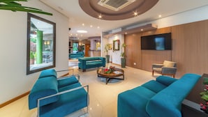A modern lobby with teal sofas, a coffee table with flowers, a TV, and a reception desk. - Hotel Laghetto Viale (Gramado)