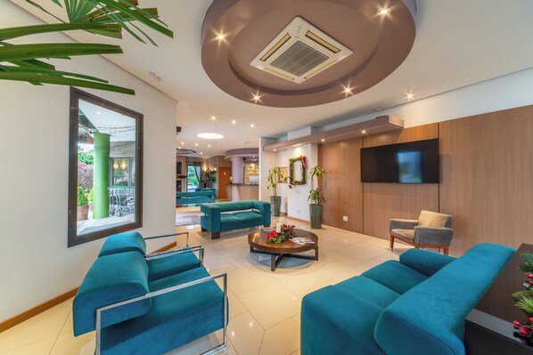 A modern lobby with teal sofas, a coffee table with flowers, a TV, and a reception desk. - Hotel Laghetto Viale (Gramado)