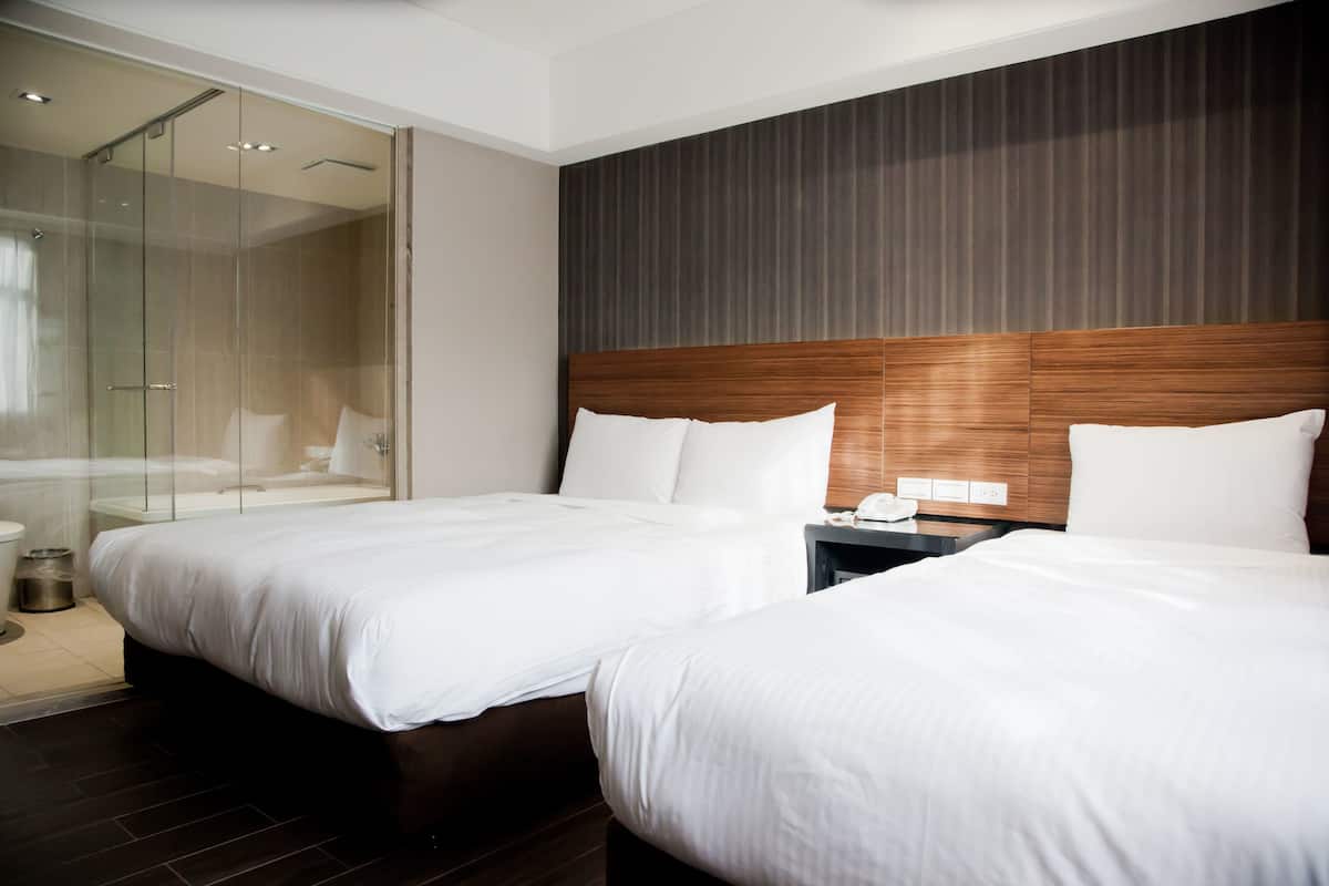 A modern hotel room with two beds, a glass shower enclosure, and a wooden headboard.