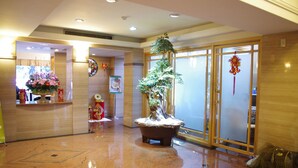 A lobby with a large potted tree, a reception desk with floral decorations, and a traditional Chinese decorative hanging. - KDM Hotel (Taipei)