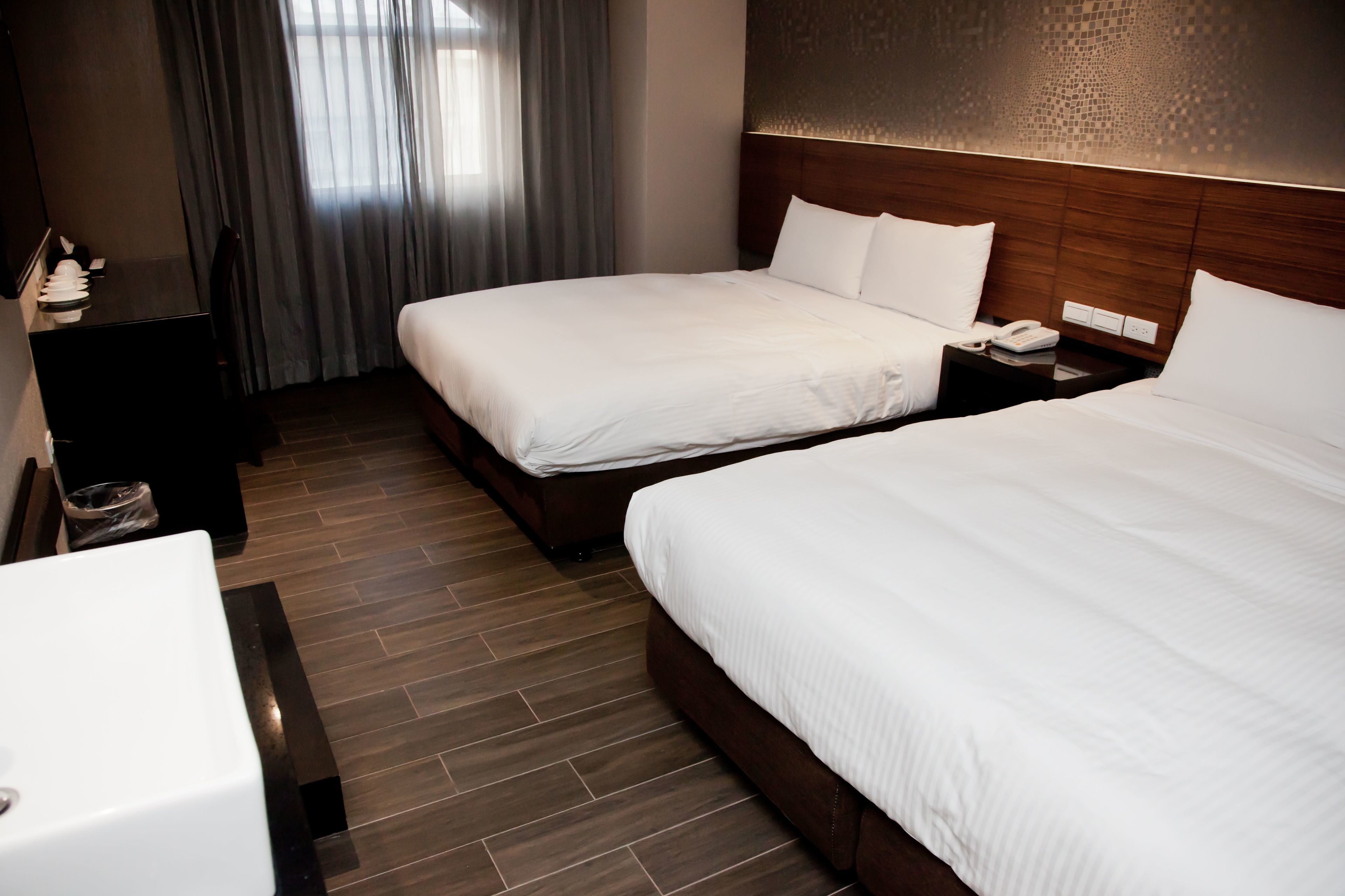 A hotel room with two beds, a wooden headboard, a bedside table with a phone, and a window with curtains.