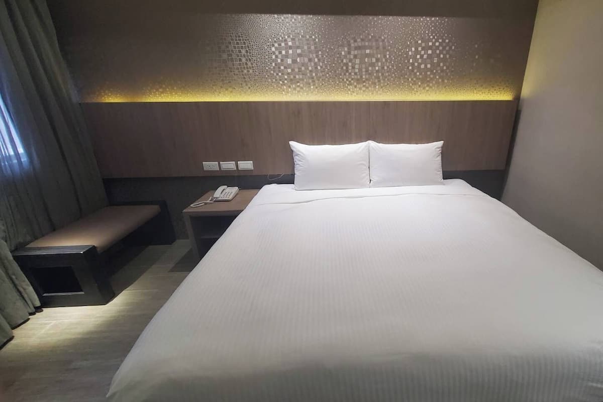 A hotel room with a large bed, a bedside table with a phone, and a bench.