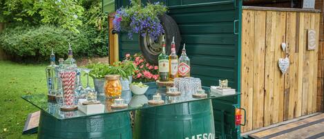 Una barra con dos barriles verdes etiquetados 'JAMESON', un neumático decorado con flores y varias botellas y vasos sobre una mesa.