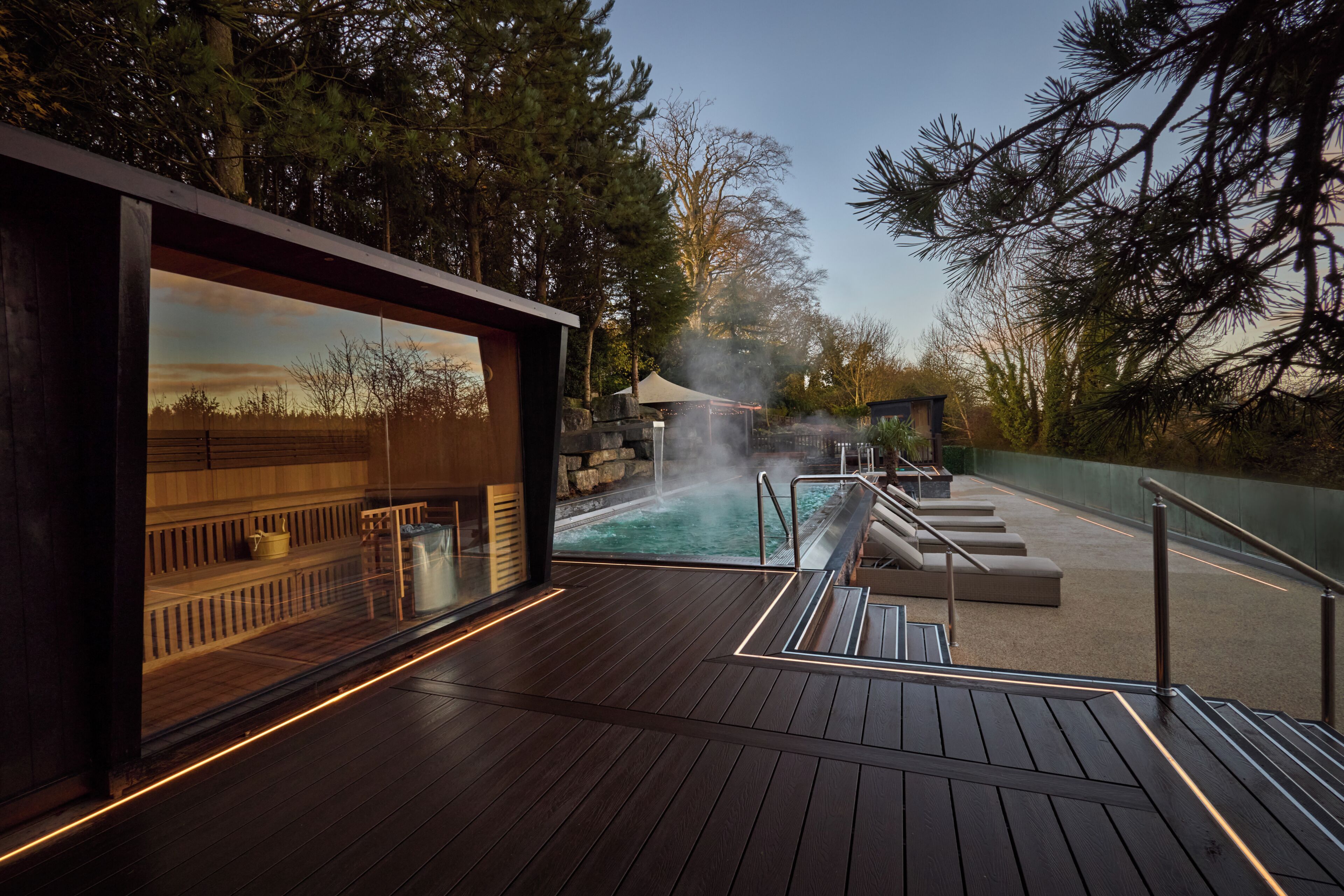A wooden deck with a pool, steam feature, and a view of trees.