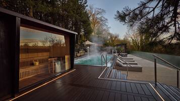 A wooden deck with a pool, steam feature, and a view of trees.