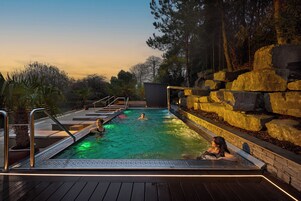 Una piscina al aire libre climatizada con cubierta de madera, rodeada de rocas y árboles.