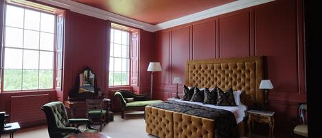 A room with a large bed, two side tables, a chair, a small table, and a mirror.