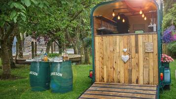 A rustic wooden structure with a ramp leading to a bar area, surrounded by greenery and barrels.