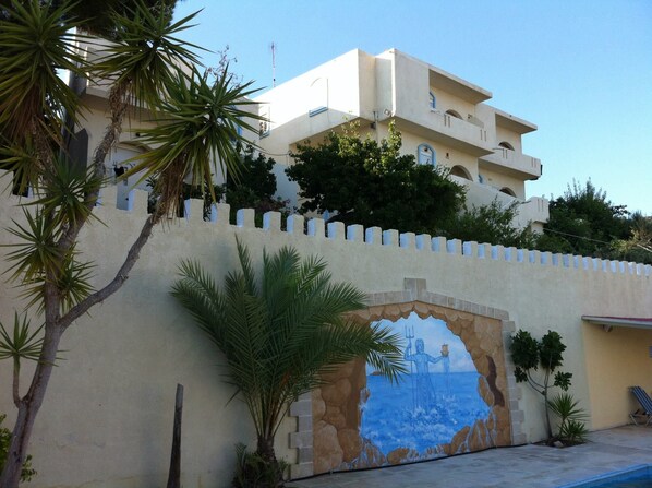A building with a mural of a sea scene on the wall, palm trees, and a clear sky. - Achlia Hotel (Ierapetra)