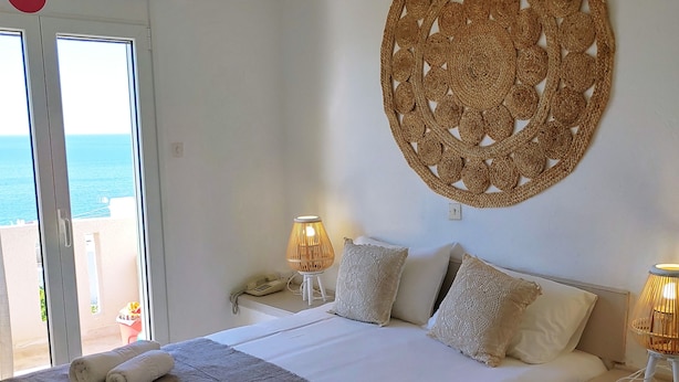 A hotel room with a bed, two bedside lamps, a woven wall hanging, a door with a red circle sign, and a view of the sea through the window. - Achlia Hotel (Ierapetra)