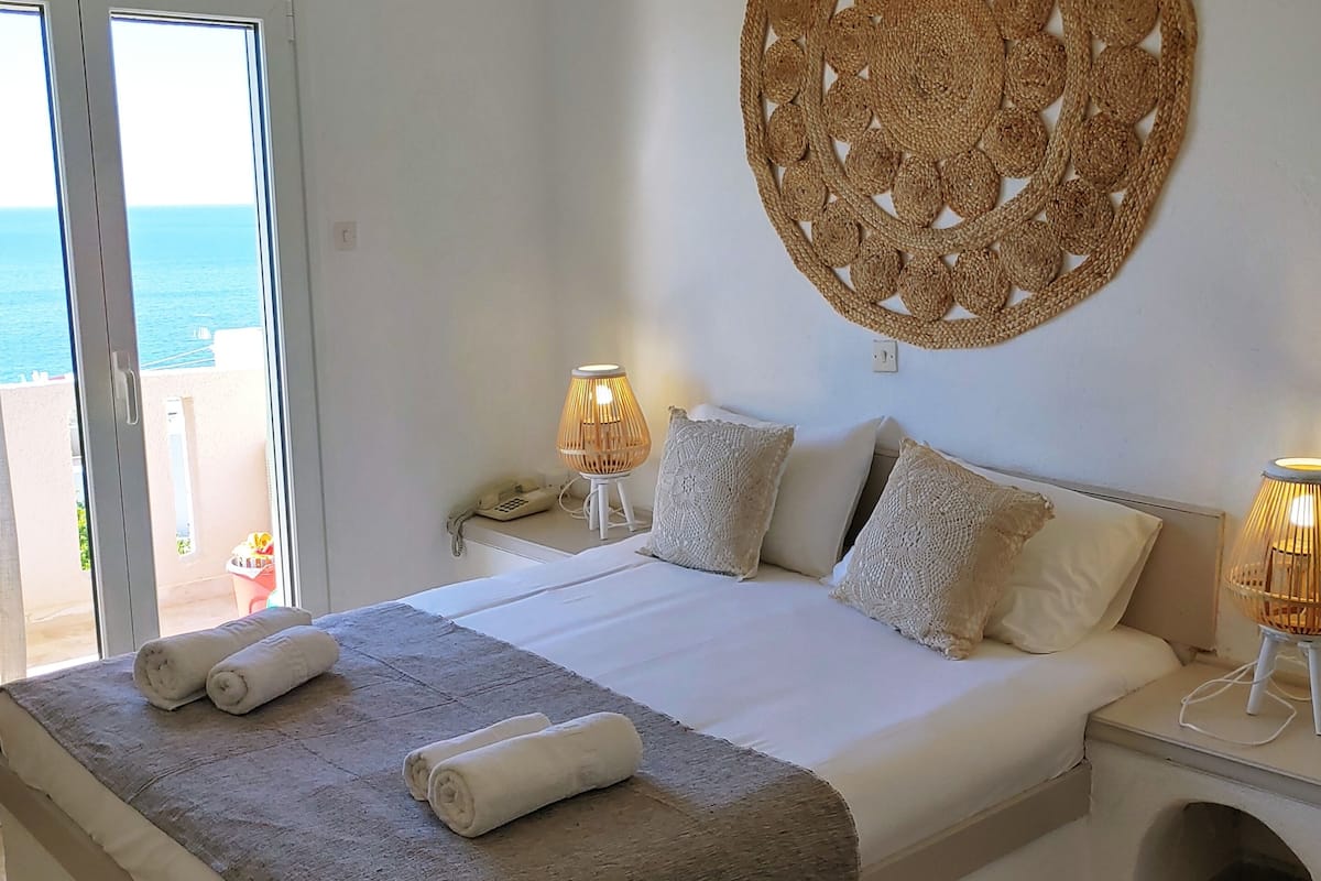 Ein Hotelzimmer mit einem Bett, zwei Nachttischlampen, einem Wandteppich, einer Tür mit einem roten Kreis-Schild und Meerblick durch das Fenster.