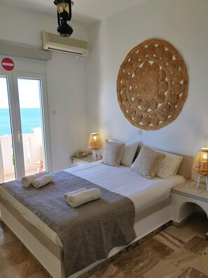 A hotel room with a bed, two bedside lamps, a woven wall hanging, a door with a red circle sign, and a view of the sea through the window.