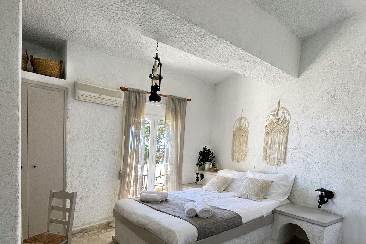 Ein Schlafzimmer mit einem großen Bett, einem hölzernen Stuhl, einem Nachttisch mit Lampe und einem Wandbehang aus Makramee.