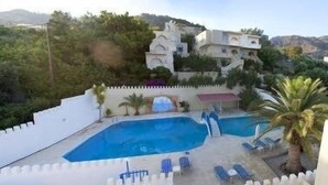 A swimming pool with lounge chairs and a palm tree, surrounded by a white fence and a building with a unique architectural style. - Achlia Hotel (Ierapetra)