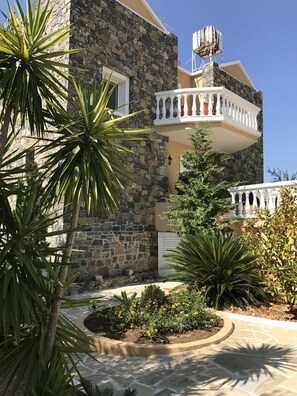 A stone building with a balcony, surrounded by tropical plants and a paved driveway. - Achlia Hotel (Ierapetra)