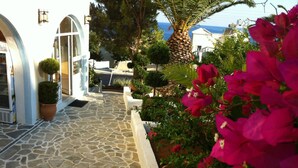A stone-paved pathway leading to a white building with arched doorways, surrounded by lush greenery and vibrant pink flowers. - Achlia Hotel (Ierapetra)