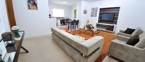 Una sala moderna con un sofá, una mesa de centro y un televisor montado en la pared. Se ve un comedor al fondo con una mesa y sillas.