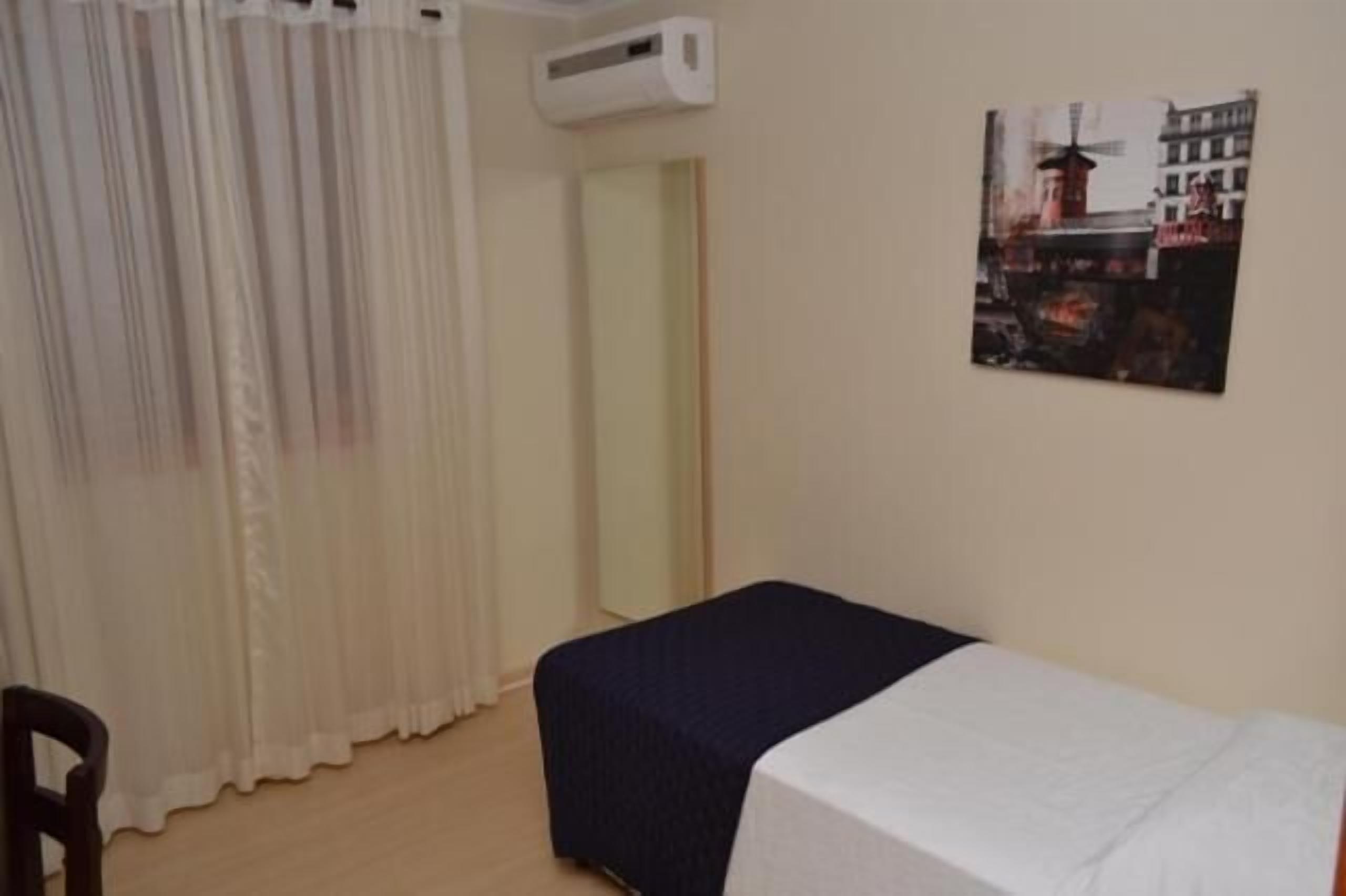 Una habitación pequeña con una cama, una silla, un ventanal con cortinas traslúcidas y un aire acondicionado de pared.