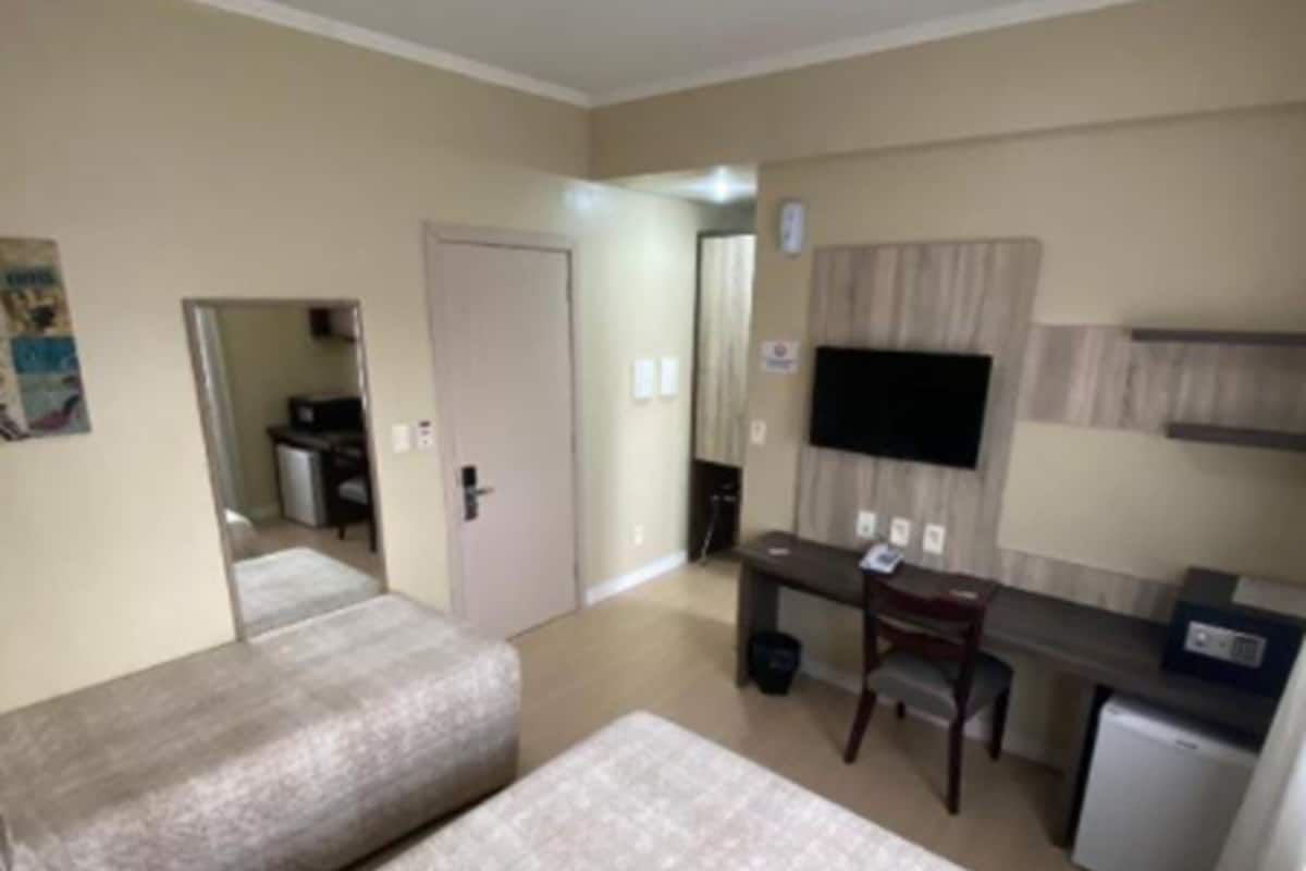 Ein Hotelzimmer mit zwei Betten, einem Schreibtisch mit Stuhl, einer kleinen Küchenzeile, einem wandmontierten Flachbildfernseher und einem Spiegel.