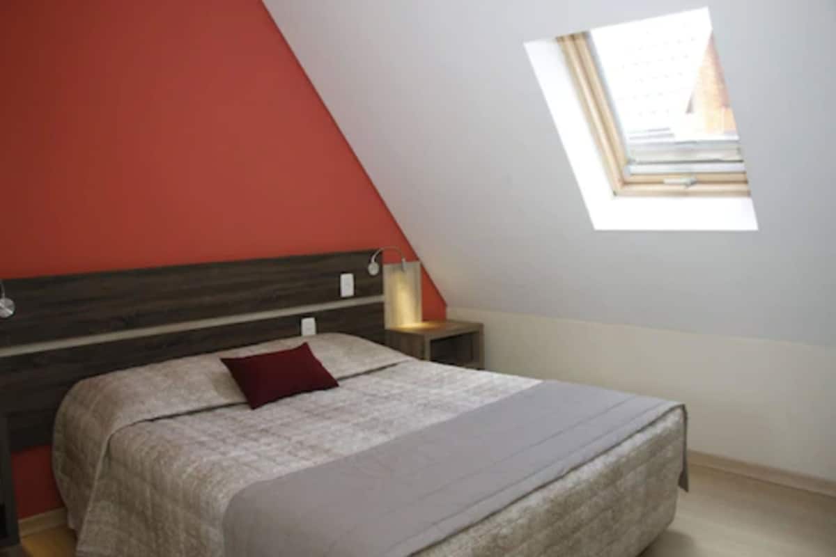 Ein Schlafzimmer mit schrägen Wänden, einem Bett mit rotem Kissen, einem Nachttisch und einem Fenster mit Blick ins Freie.