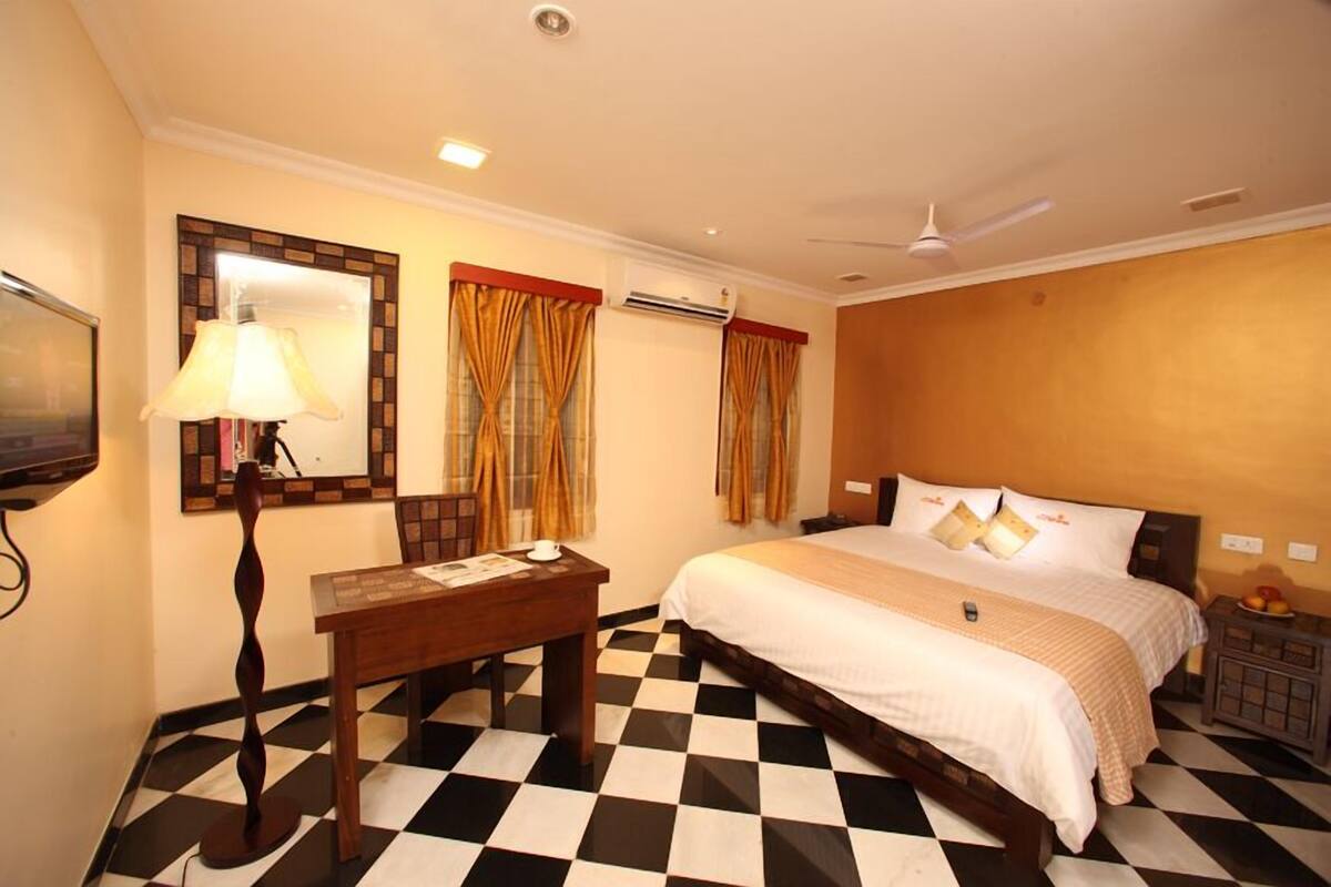 A hotel room with a checkered floor, a bed with white bedding, a desk with a lamp, and a mirror with a decorative frame.