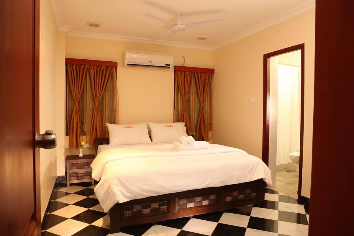 A hotel room with a checkered floor, a bed with white linens, curtains, an air conditioning unit, and a ceiling fan.