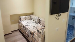 Um quarto pequeno com um sofá coberto por padrão de jornal, uma televisão de tela plana fixada na parede e um banheiro visível através de uma porta de vidro.