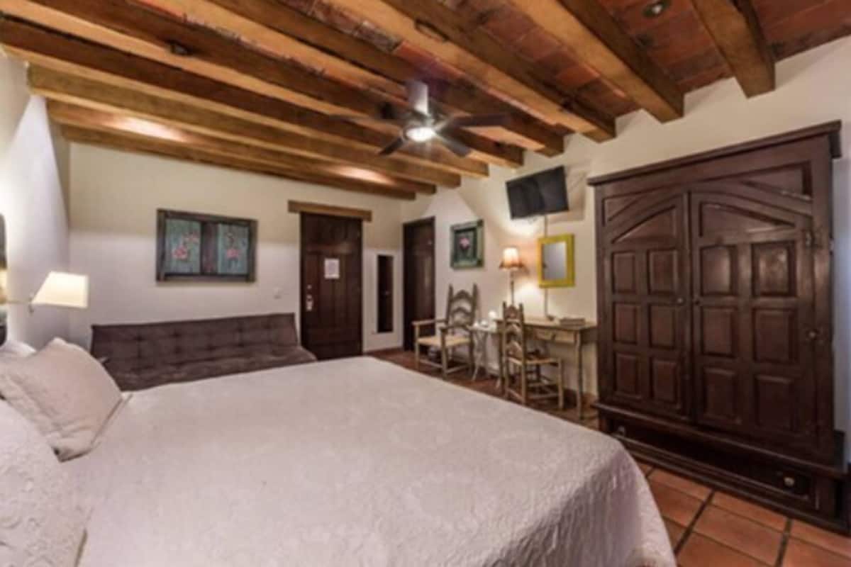 A bedroom with a large bed, wooden ceiling beams, a desk with chairs, and a wardrobe.