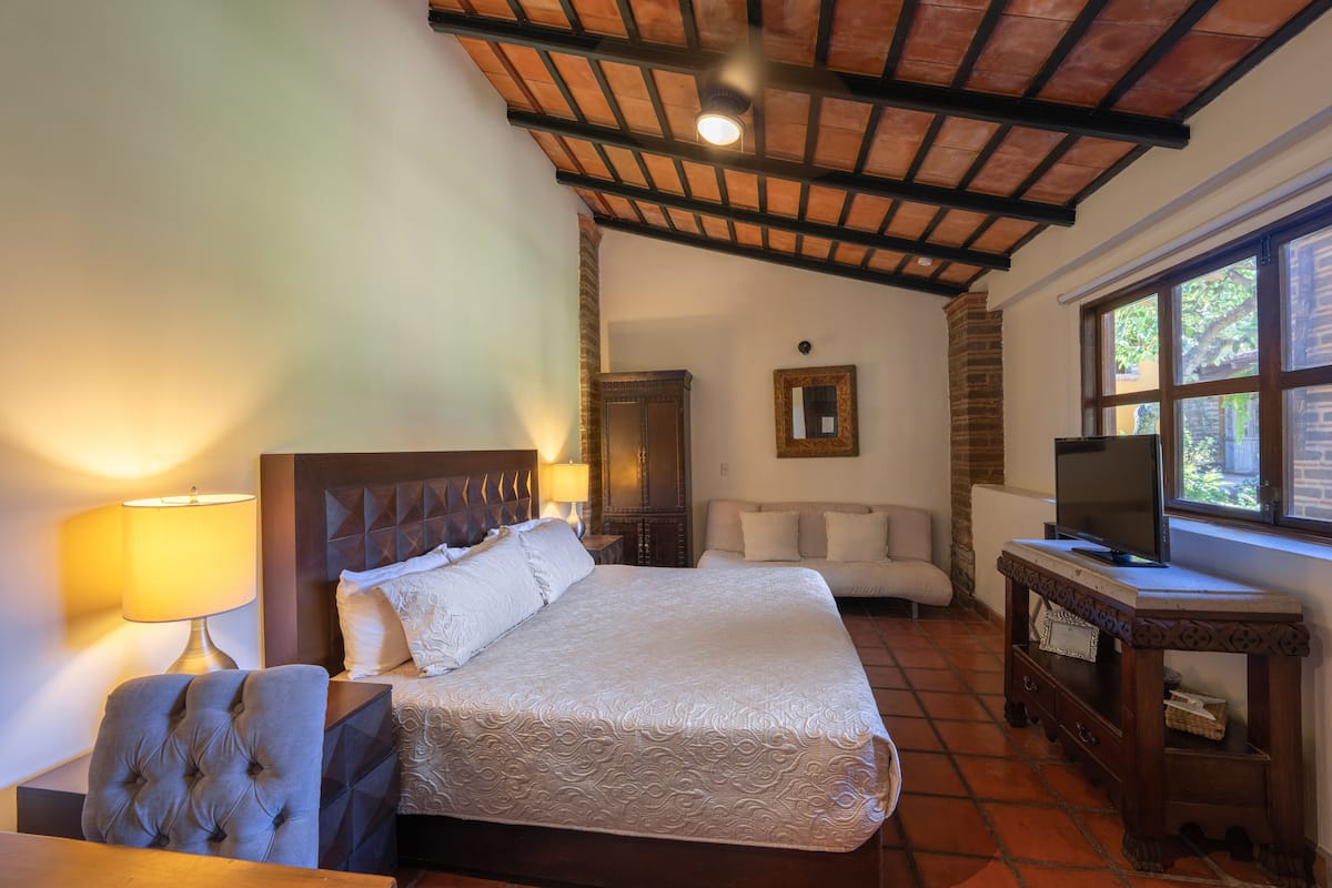 A hotel room with a large bed, a TV on a wooden table, a lamp, and a window with a view of greenery.